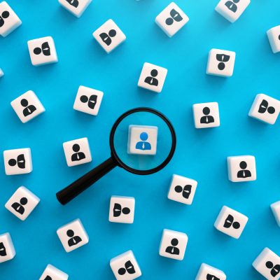 Magnifying glass focus on white square with blue figure in a group of white squares with black figure against a blue background.